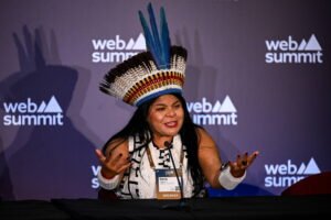 Mulher indígena com cocar colorido e pulseiras brancas fala em mesa de conferência no evento Web Summit, com fundo roxo e logo do evento.