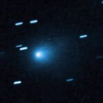 Cometa azul brilhante com núcleo central luminoso e cauda difusa se estendendo para a esquerda, cercado por estrelas com traços de movimento em fundo escuro.