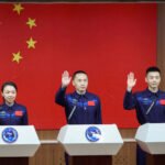 Três astronautas chineses em uniformes azuis com bandeira da China no peito levantam a mão direita em gesto de juramento diante de microfones. Ao fundo, grande bandeira da China vermelha com estrelas amarelas.