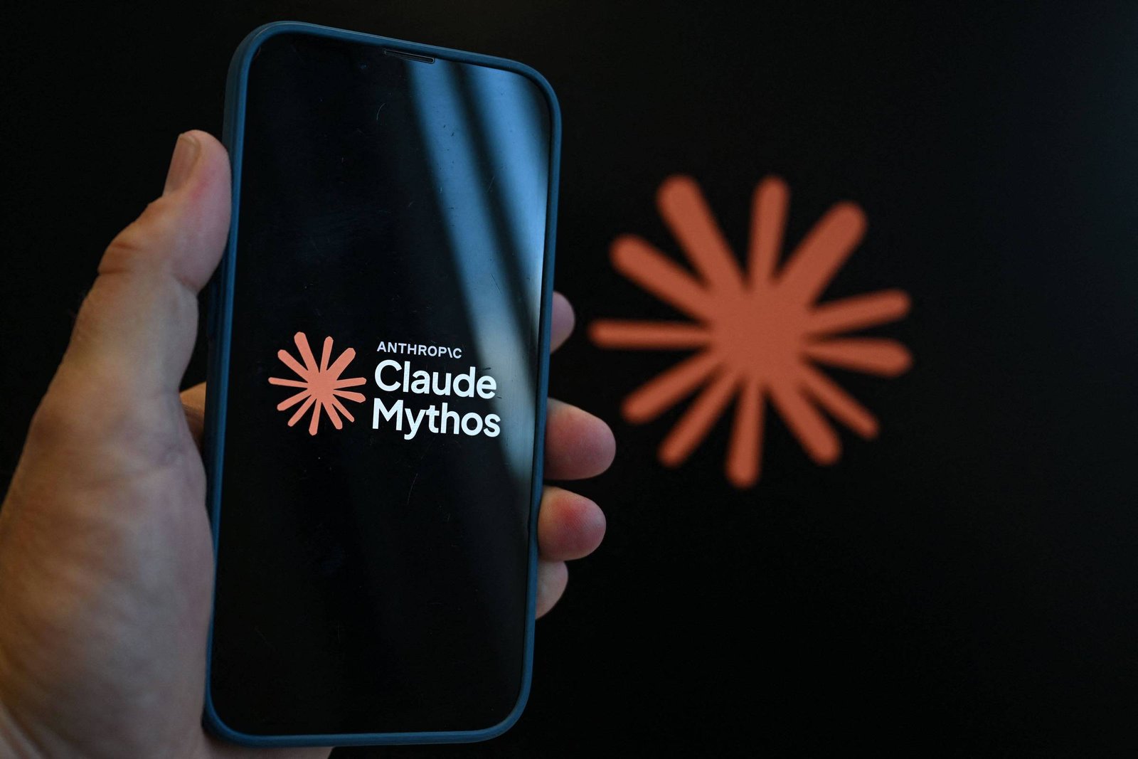 Mão segurando smartphone com tela preta que mostra o logo do Claude Mythos em branco e laranja. Ao fundo, símbolo laranja desfocado em fundo preto.