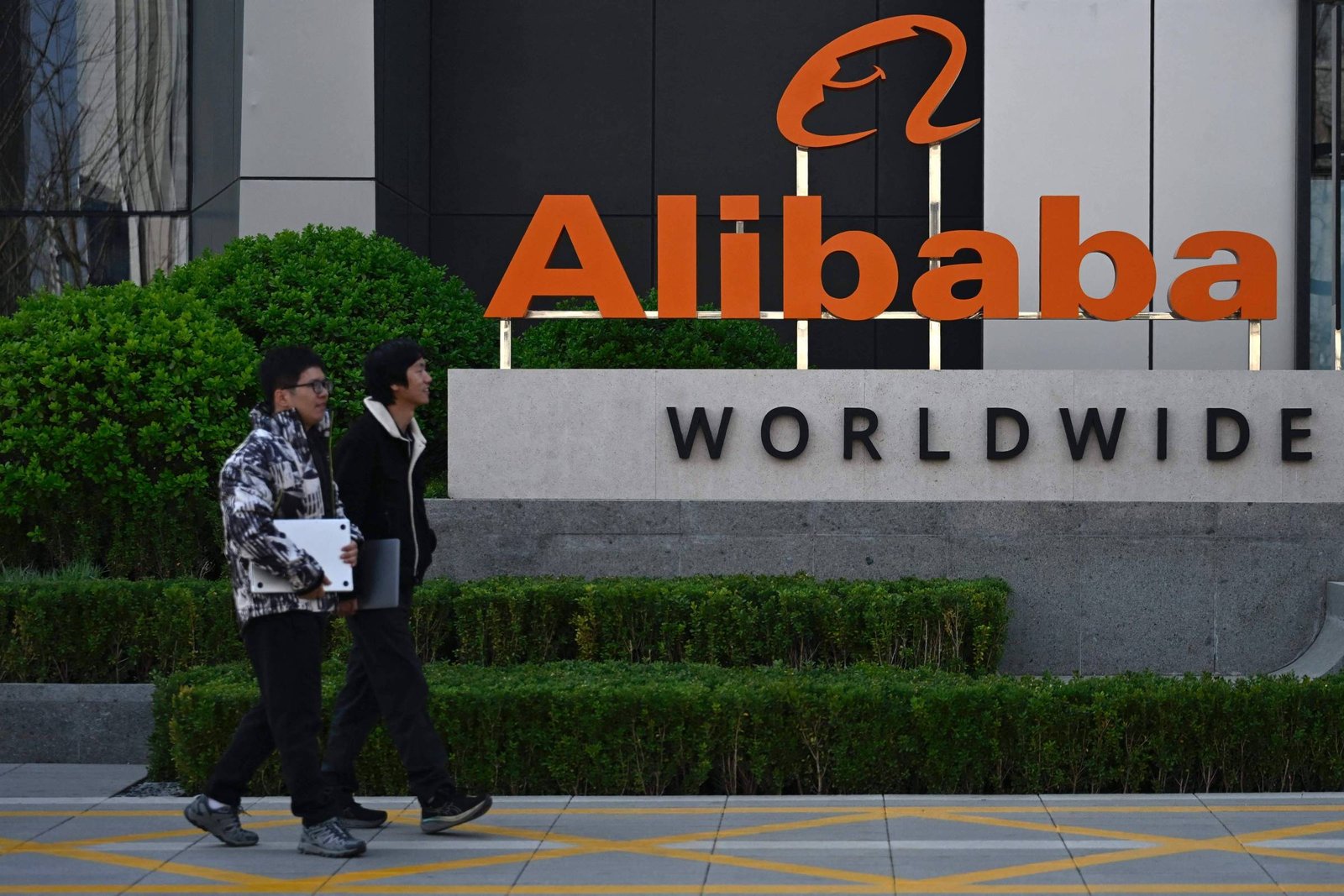 Duas pessoas caminham em frente à placa da Alibaba Worldwide, com o logotipo laranja da empresa em destaque. A área externa tem arbustos verdes e um prédio ao fundo.