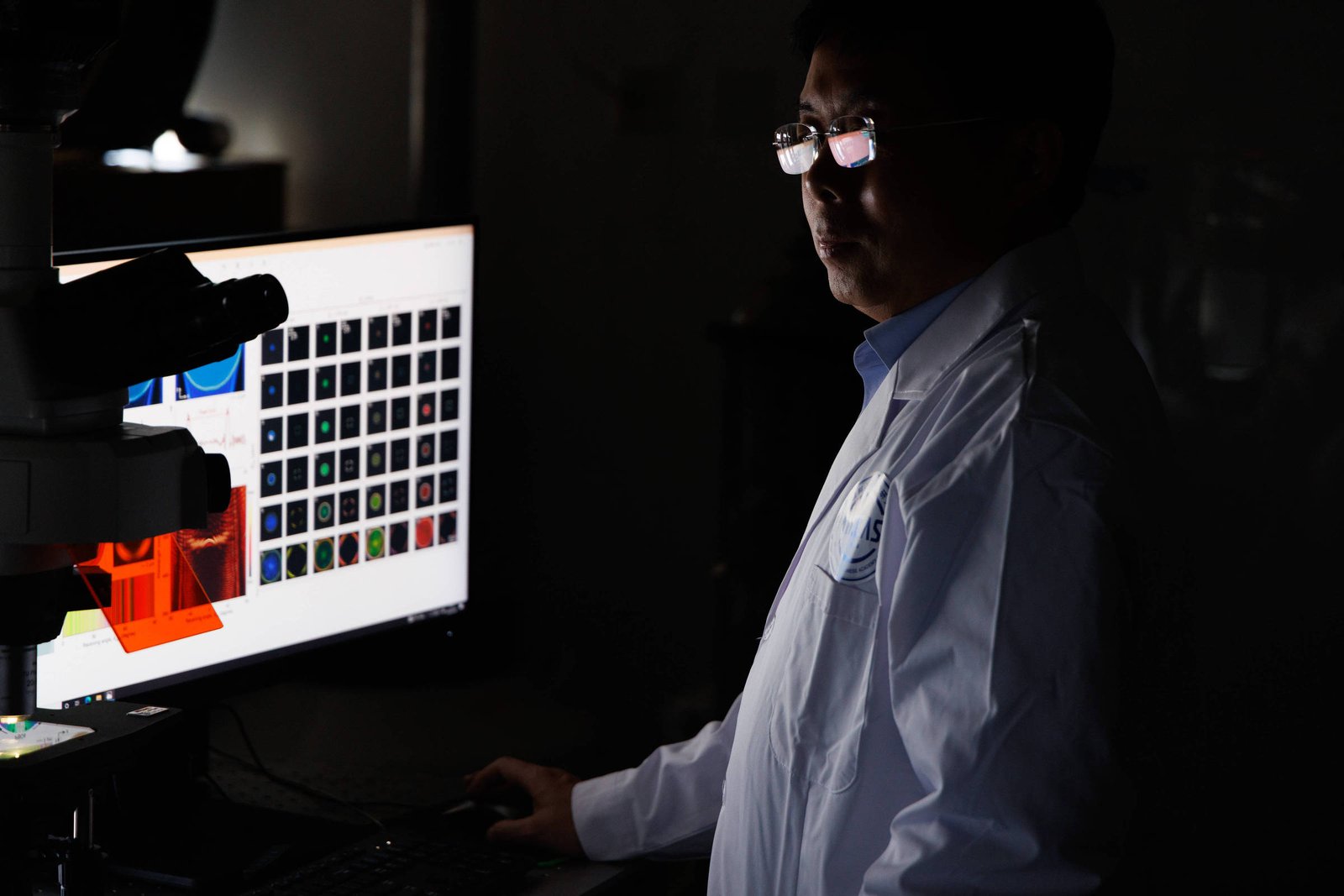 Pesquisador de jaleco branco observa imagens coloridas em tela de computador em ambiente escuro, com microscópio ao lado.