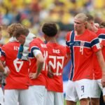 Seis jogadores da seleção norueguesa de futebol reunidos no campo durante intervalo. Eles vestem uniformes vermelhos com detalhes em azul e branco, e shorts brancos. Um jogador de cabelo loiro comprido destaca-se no centro da imagem.