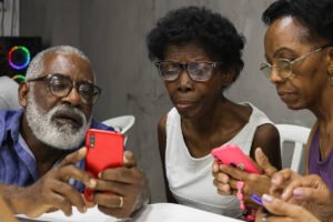 Três pessoas idosas sentadas lado a lado em cadeiras brancas, cada uma segurando um smartphone colorido. O homem à esquerda, com barba branca e óculos, mostra algo no celular vermelho para as duas mulheres ao lado, que também usam óculos e observam atentamente as telas.