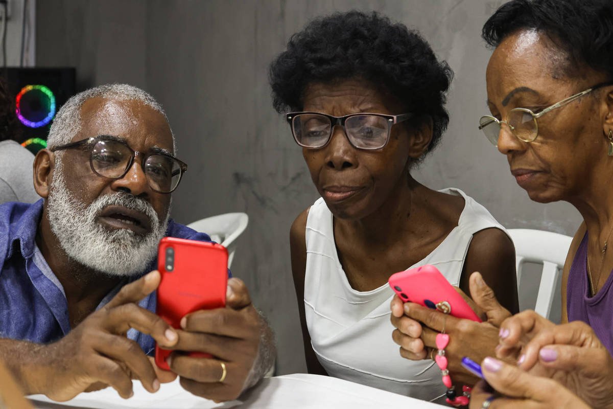 Três pessoas idosas sentadas lado a lado em cadeiras brancas, cada uma segurando um smartphone colorido. O homem à esquerda, com barba branca e óculos, mostra algo no celular vermelho para as duas mulheres ao lado, que também usam óculos e observam atentamente as telas.