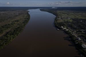 Vista aérea de um rio largo com vegetação densa à esquerda e área urbana à direita, sob céu parcialmente nublado.