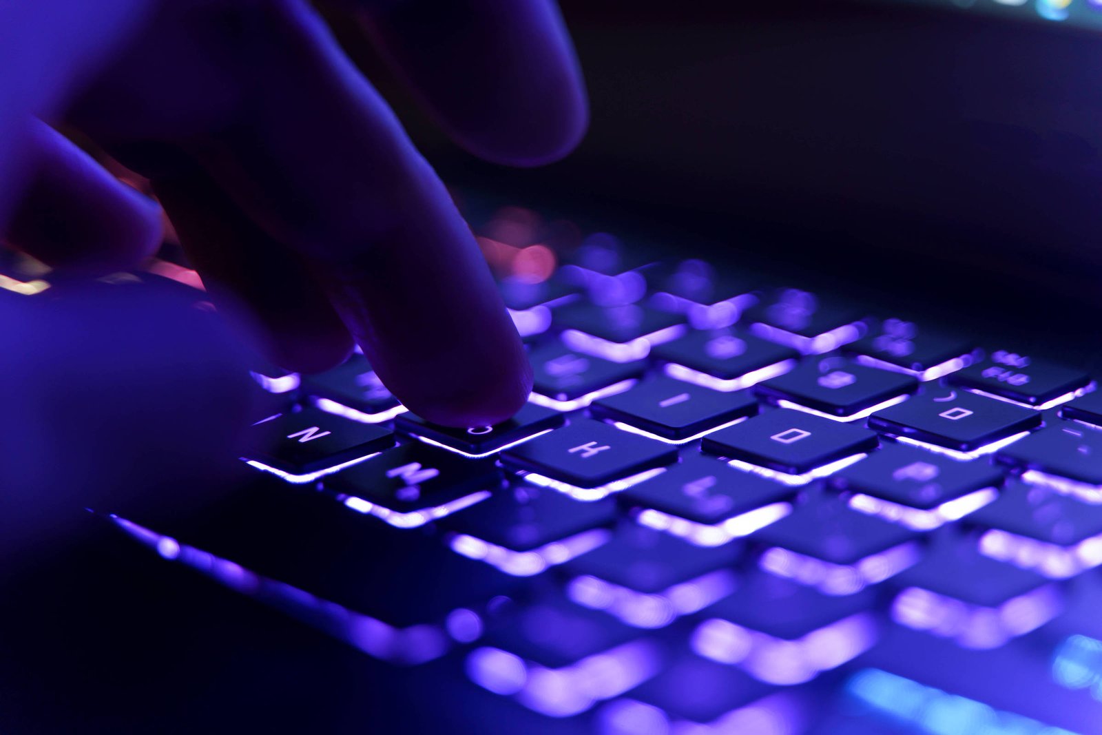 Dedos pressionam teclas iluminadas em teclado de laptop com luz roxa em ambiente escuro.