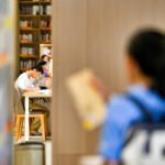 Dois estudantes sentados em mesa branca em corredor de biblioteca, focados na leitura. Prateleiras com livros ao fundo e outros estudantes em primeiro plano desfocados.