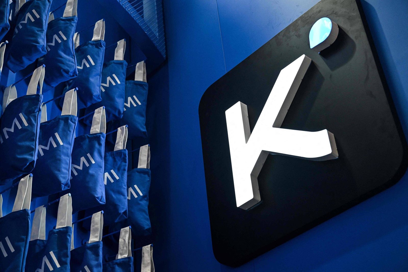 Logotipo branco da Kovi em fundo preto fixado em parede azul. Ao lado, várias mochilas azuis com a palavra