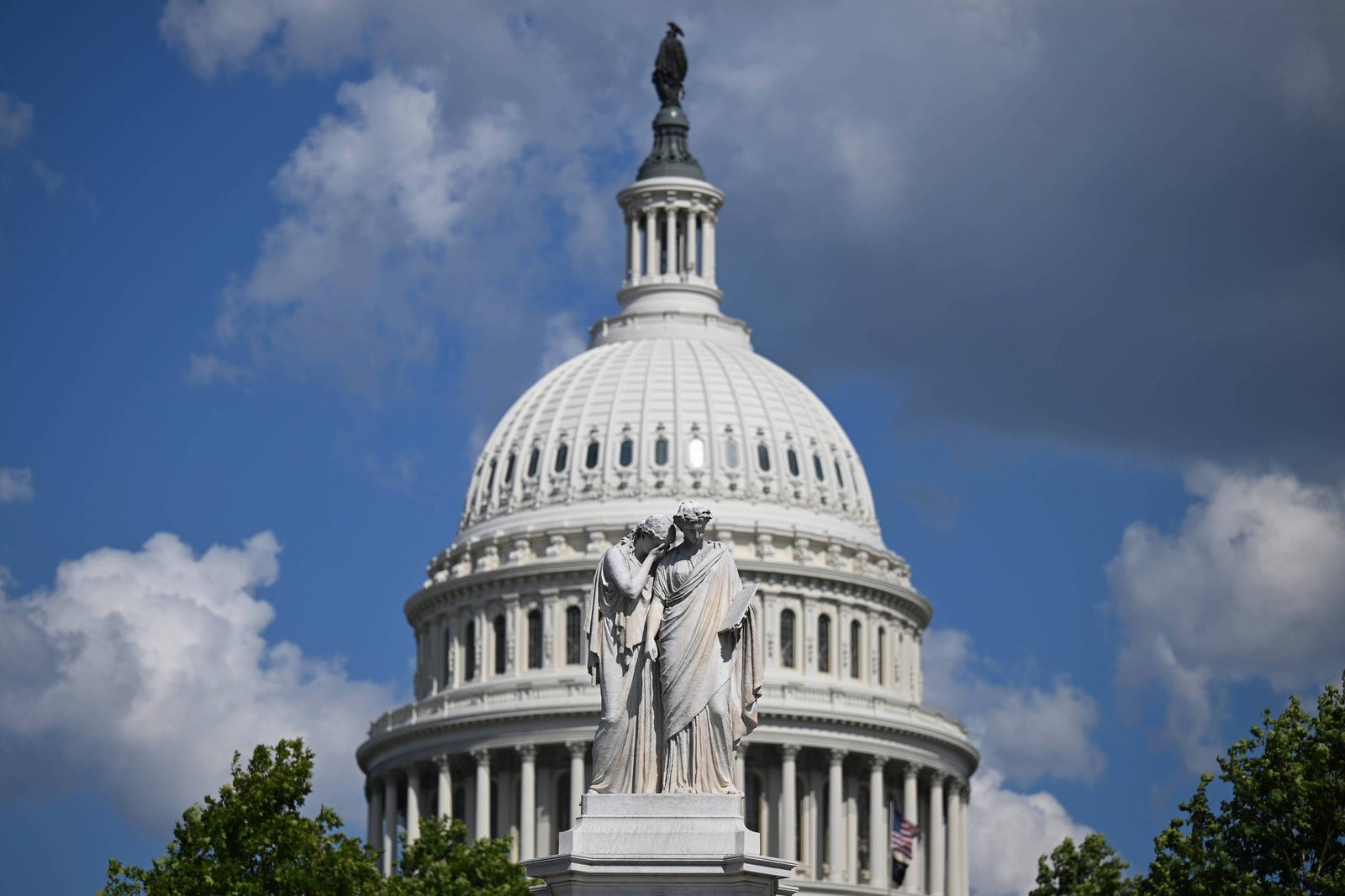 Cúpula branca do Capitólio dos Estados Unidos sob céu parcialmente nublado. Estátua de mármore com duas figuras humanas em primeiro plano, cercada por árvores verdes.