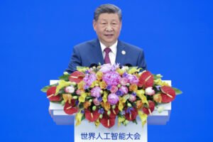Homem em terno azul e gravata vermelha fala em púlpito branco decorado com arranjo floral colorido. Fundo azul sólido com logotipo e texto em chinês da Conferência Mundial de Inteligência Artificial.