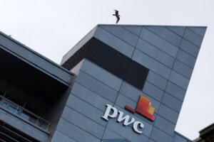 Prédio moderno com fachada cinza e logo colorida da PwC em destaque. Gaivota voa acima do topo do edifício sob céu nublado.