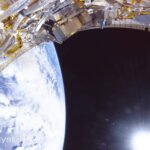 Parte de um módulo espacial com componentes metálicos e cabos visível no canto superior esquerdo. Abaixo, a Terra aparece com nuvens brancas sobre o oceano azul. O espaço escuro e o sol brilhante estão à direita.