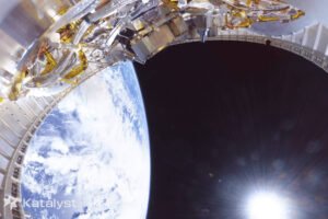 Parte de um módulo espacial com componentes metálicos e cabos visível no canto superior esquerdo. Abaixo, a Terra aparece com nuvens brancas sobre o oceano azul. O espaço escuro e o sol brilhante estão à direita.