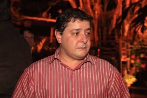 Homem de meia-idade veste camisa listrada vermelha e branca, olhando para o lado direito. Fundo desfocado com iluminação laranja e elementos que sugerem ambiente interno ou festa.