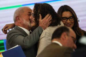 Um homem e uma mulher se abraçam e um deles beija o rosto do outro em ambiente interno com fundo desfocado. Uma mulher de óculos sorri ao fundo, e outra pessoa aparece parcialmente em primeiro plano.