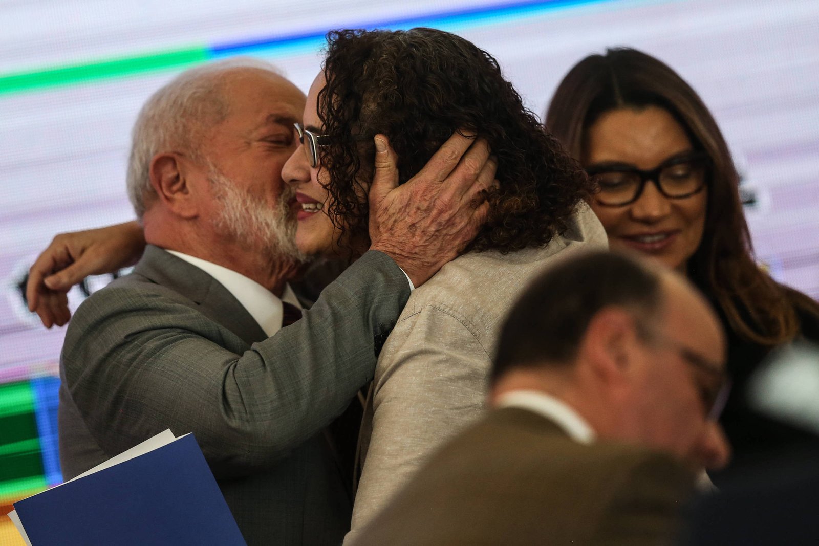 Um homem e uma mulher se abraçam e um deles beija o rosto do outro em ambiente interno com fundo desfocado. Uma mulher de óculos sorri ao fundo, e outra pessoa aparece parcialmente em primeiro plano.