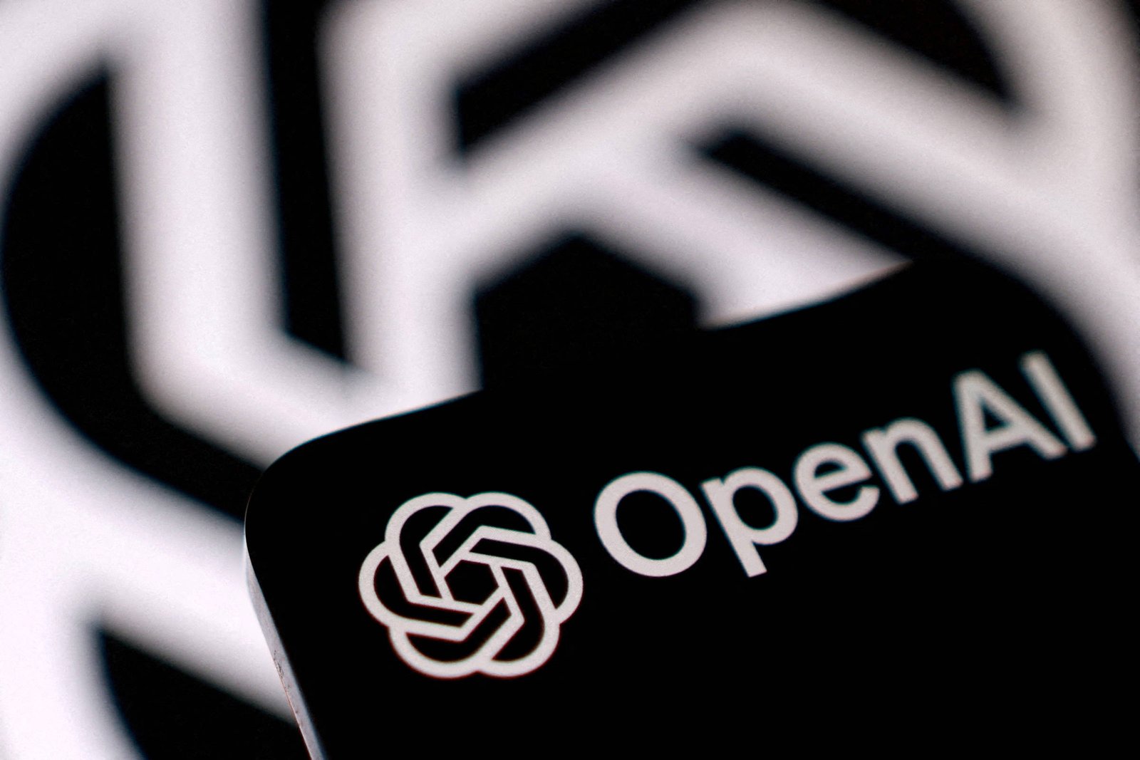 Logotipo branco da OpenAI com texto ao lado exibido em tela escura de dispositivo móvel. Fundo desfocado mostra padrão semelhante ao logotipo em preto e branco.