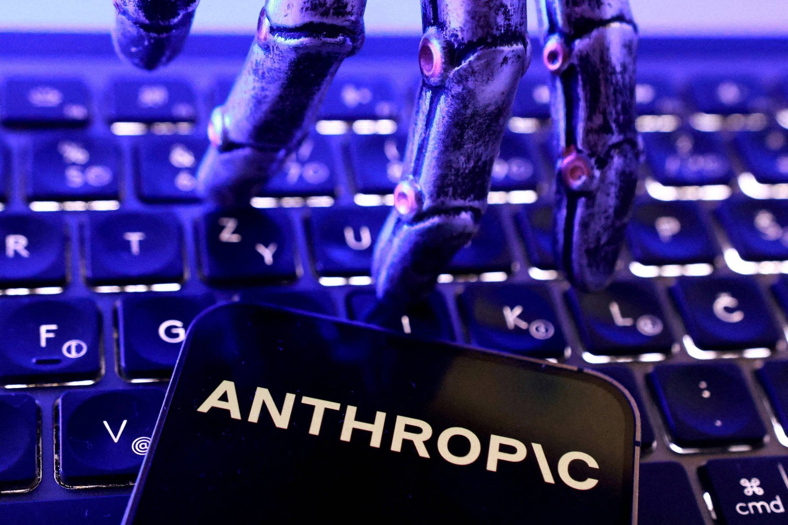 Duas mãos metálicas robóticas posicionadas sobre teclado de computador iluminado em azul. Na frente, celular exibe a palavra