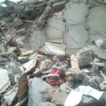 Escombros de concreto, metal e madeira de um edifício desabado. Fios de aço expostos e pedaços de paredes quebradas estão visíveis. Documentos e objetos pessoais espalhados entre os destroços.