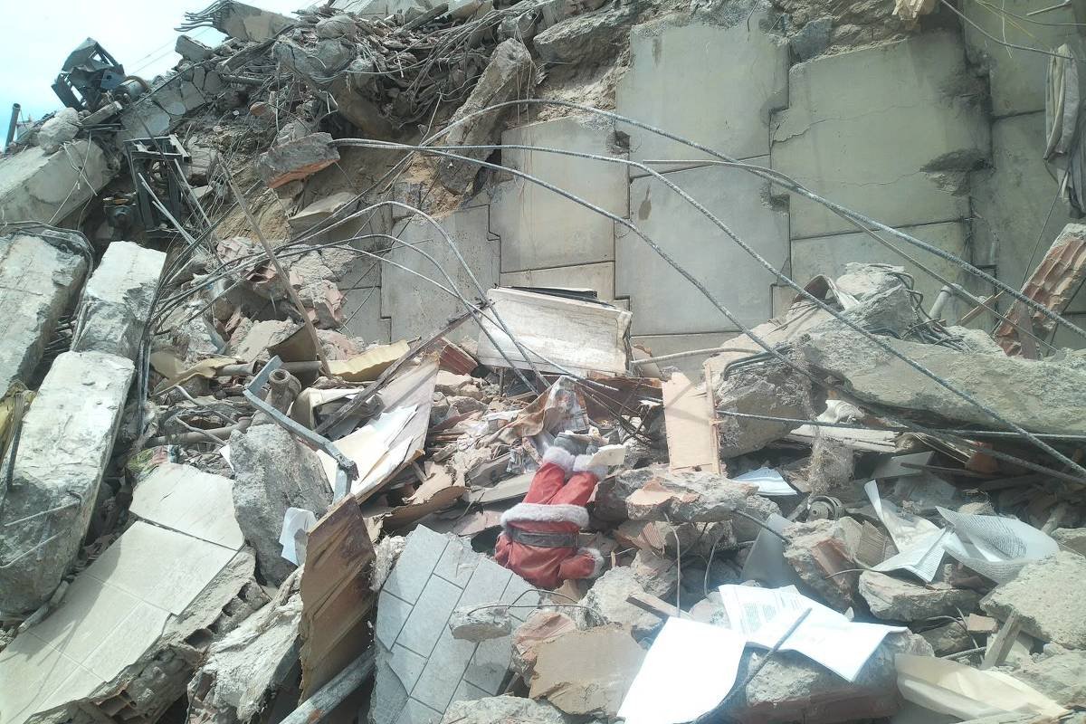 Escombros de concreto, metal e madeira de um edifício desabado. Fios de aço expostos e pedaços de paredes quebradas estão visíveis. Documentos e objetos pessoais espalhados entre os destroços.