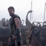 Homem vestido com armadura de couro preta e espada na cintura está em primeiro plano, olhando para a câmera. Ao fundo, grupo de pessoas e um navio viking com velas e mastros visíveis em ambiente enevoado.