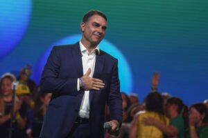 Homem de terno azul e camisa branca segura microfone e coloca a mão no peito em palco com fundo azul. Público desfocado ao fundo, alguns com roupas verdes e amarelas.