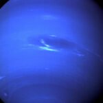 Imagem do planeta Netuno mostrando sua coloração azul intensa com faixas claras e uma mancha escura visível no hemisfério superior direito. O fundo é completamente preto, destacando o planeta.