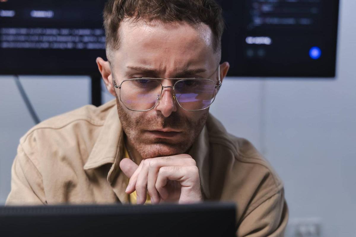 Homem de óculos e camisa bege olha atentamente para a tela de um computador, com a mão no queixo, em ambiente interno com telas ao fundo.