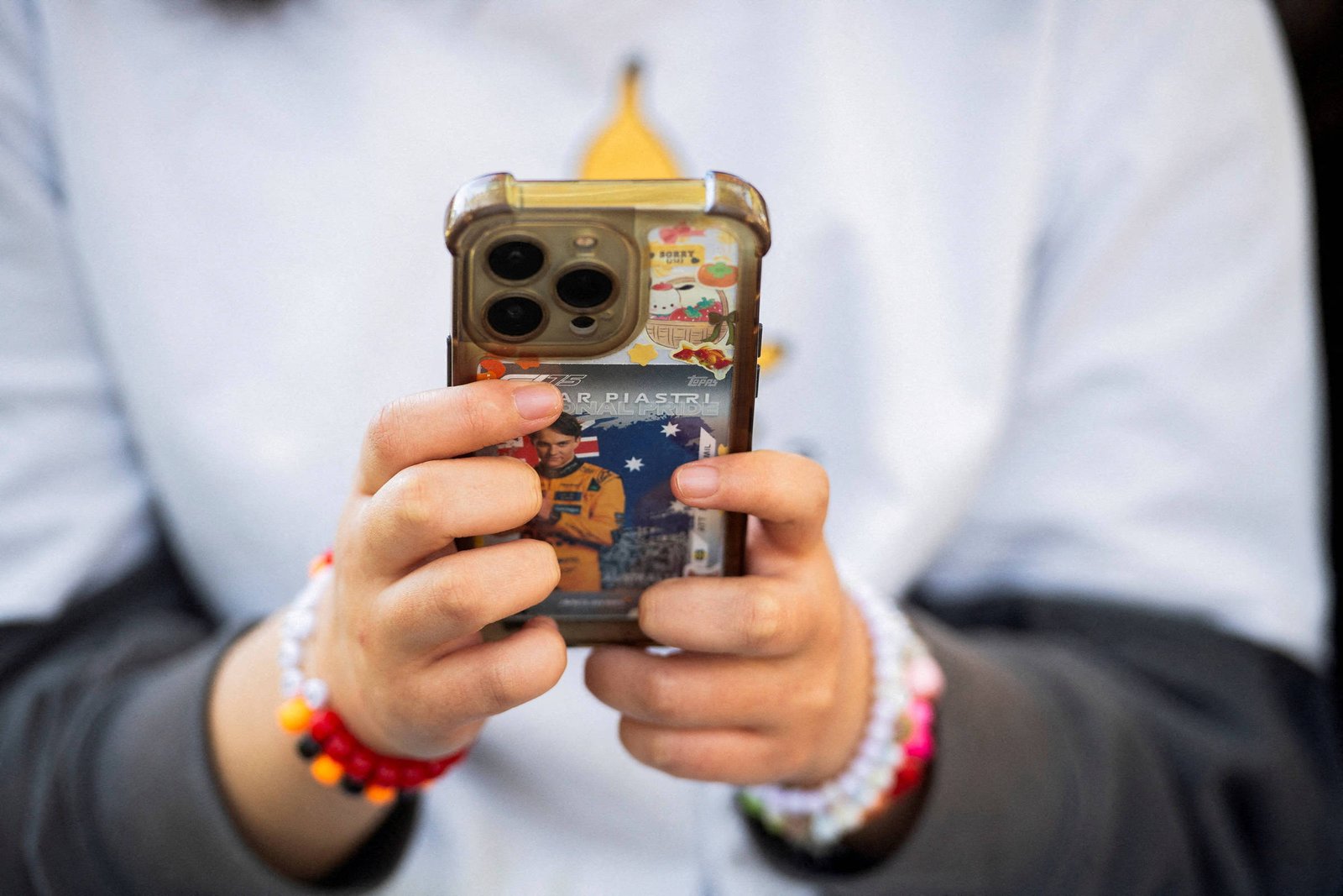 Mãos seguram celular com capa transparente contendo imagens e cartões, incluindo figura de astronauta e bandeira da Austrália. Pessoa veste camiseta branca e pulseiras coloridas.