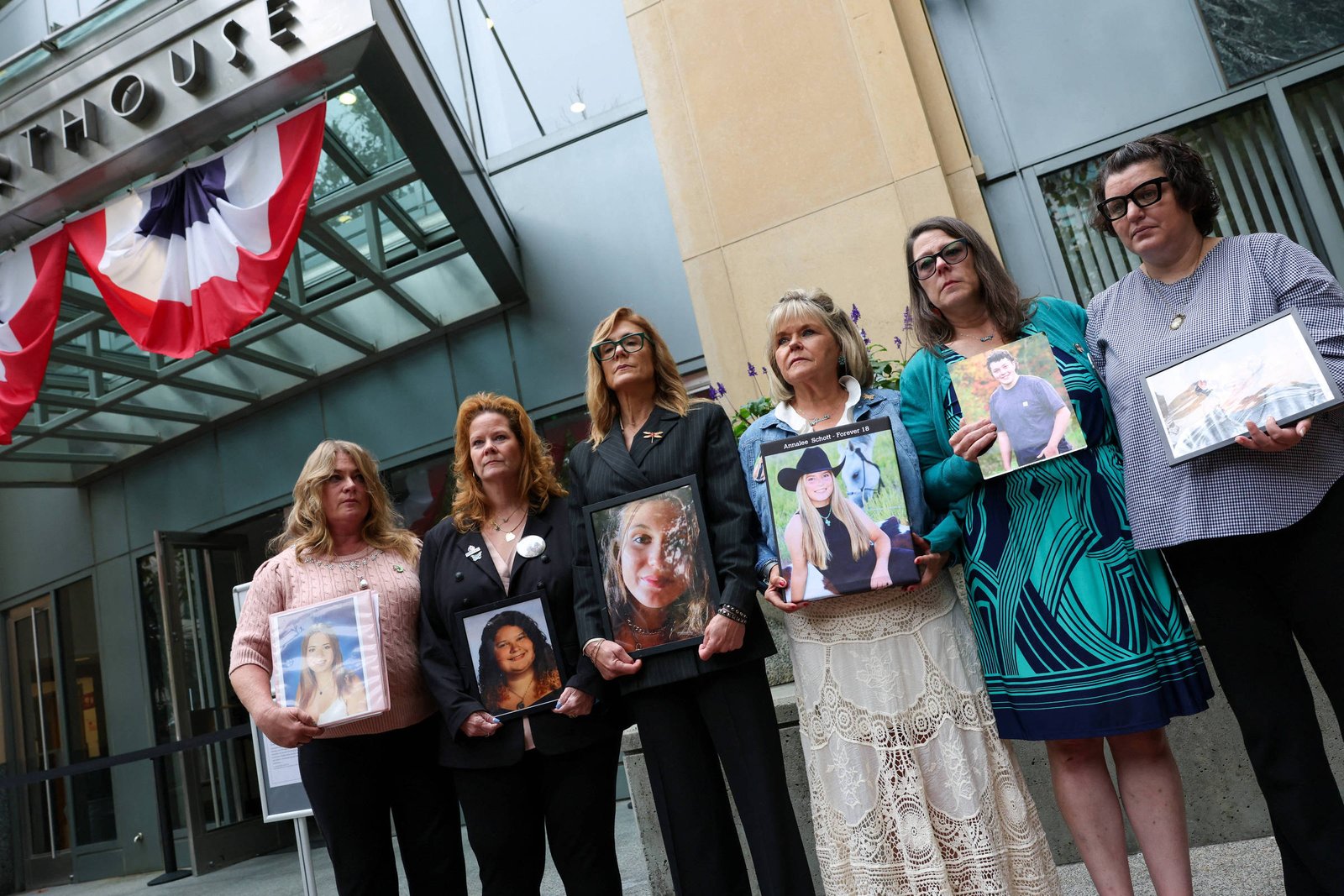 Seis mulheres em pé seguram retratos de pessoas, em frente à entrada de um tribunal decorado com bandeira vermelha, branca e azul. Elas estão alinhadas, olhando para frente, vestindo roupas formais e casuais.