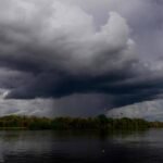 Nuvem escura e densa de tempestade cobre o céu acima de um rio calmo, com vegetação densa na margem ao fundo.