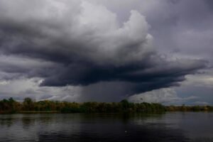 Nuvem escura e densa de tempestade cobre o céu acima de um rio calmo, com vegetação densa na margem ao fundo.