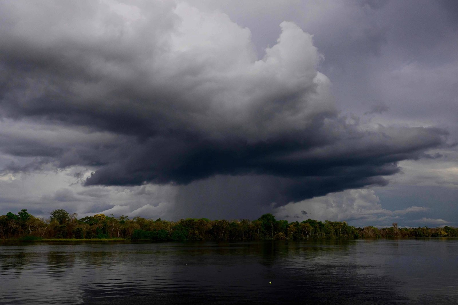 Nuvem escura e densa de tempestade cobre o céu acima de um rio calmo, com vegetação densa na margem ao fundo.
