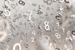 Diversos números metálicos tridimensionais em tons prateados flutuam em um espaço indefinido com fundo cinza claro desfocado.