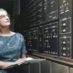 Mulher de cabelos grisalhos e óculos observa painel de controle com múltiplos botões e indicadores. Ela segura caderno aberto, em ambiente interno com iluminação baixa.