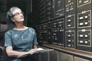 Mulher de cabelos grisalhos e óculos observa painel de controle com múltiplos botões e indicadores. Ela segura caderno aberto, em ambiente interno com iluminação baixa.