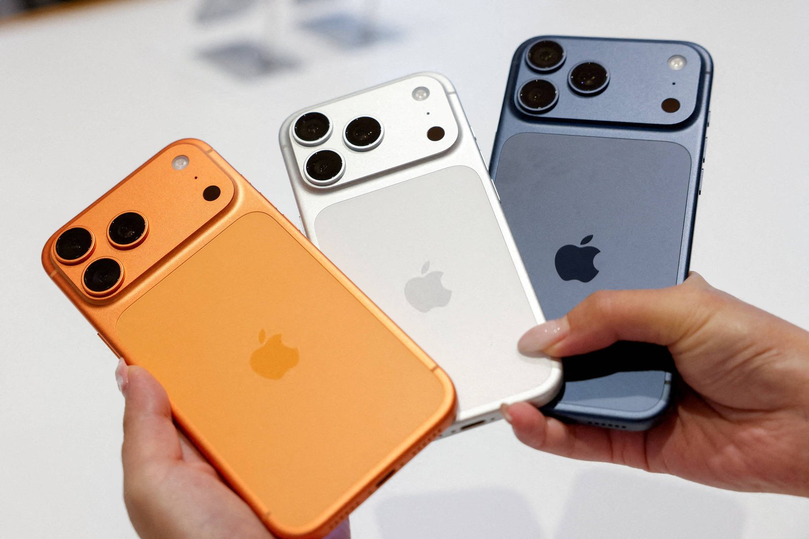 Três smartphones Apple são segurados por duas mãos, exibindo as traseiras nas cores laranja, branca e azul escuro. Cada aparelho tem três câmeras traseiras e o logo da Apple centralizado.