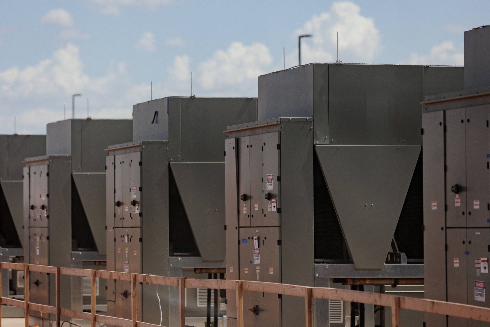 Várias unidades grandes de ar-condicionado industriais estão alinhadas em um telhado sob céu azul com poucas nuvens. Cada unidade é metálica, retangular, com painéis e aberturas visíveis, e está instalada atrás de uma grade de proteção laranja.