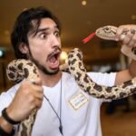 Homem de camiseta branca segura uma cobra com padrão marrom e bege enrolada em seus braços, olhando para o réptil com a boca aberta e olhos arregalados. A cobra mostra a língua vermelha bifurcada para fora. Ambiente interno com iluminação quente ao fundo.