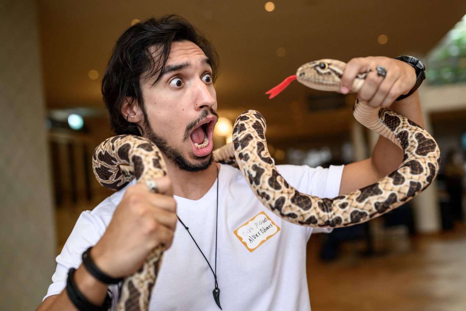 Homem de camiseta branca segura uma cobra com padrão marrom e bege enrolada em seus braços, olhando para o réptil com a boca aberta e olhos arregalados. A cobra mostra a língua vermelha bifurcada para fora. Ambiente interno com iluminação quente ao fundo.