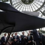 Modelo preto de aeronave futurista em exibição sob estrutura de vidro. Grupo de homens de terno observa e discute ao redor da peça.