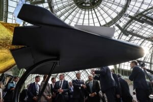 Modelo preto de aeronave futurista em exibição sob estrutura de vidro. Grupo de homens de terno observa e discute ao redor da peça.