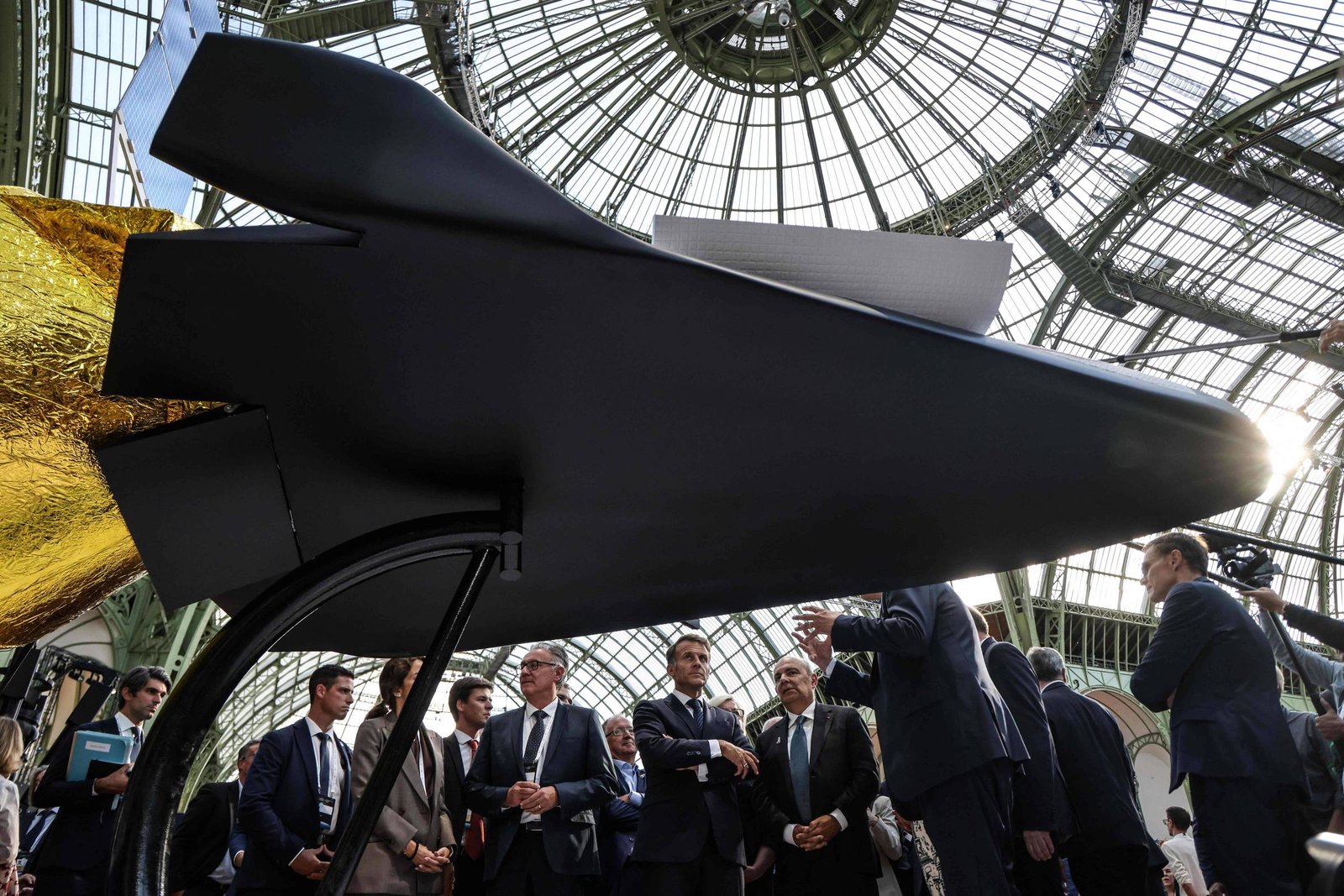 Modelo preto de aeronave futurista em exibição sob estrutura de vidro. Grupo de homens de terno observa e discute ao redor da peça.