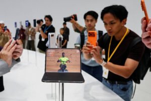 Várias pessoas tiram fotos de um smartphone dobrável exibido em pedestal branco. O aparelho mostra vídeo de homem em bicicleta, e o ambiente é iluminado e minimalista.