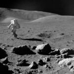 Astronauta em traje espacial completo caminha sobre terreno rochoso e irregular da Lua, com colinas suaves ao fundo e céu negro sem estrelas.