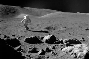 Astronauta em traje espacial completo caminha sobre terreno rochoso e irregular da Lua, com colinas suaves ao fundo e céu negro sem estrelas.