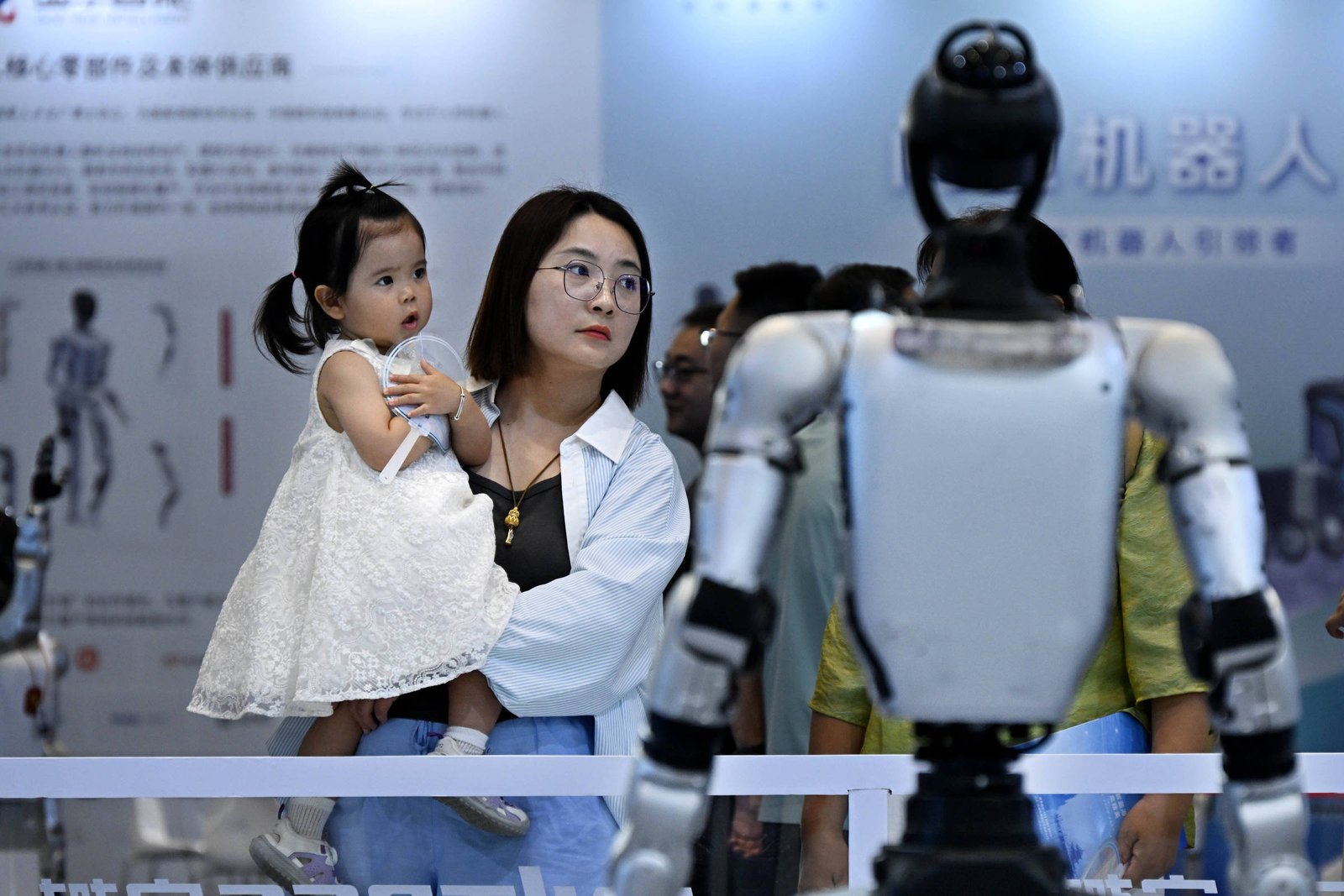 Mulher segura criança vestida de branco enquanto ambas olham para robô humanoide em exposição. Ambiente interno com outras pessoas ao fundo e painéis informativos.