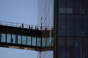 Sete pessoas caminham sobre passarela envidraçada que conecta dois prédios altos de vidro sob céu azul. Reflexos das pessoas aparecem na fachada do prédio à direita.