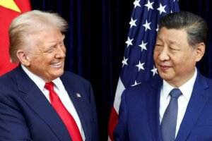 Dois homens em trajes formais, um com terno azul escuro, camisa branca e gravata vermelha, o outro com terno azul, camisa branca e gravata azul, estão lado a lado. Ao fundo, bandeiras dos Estados Unidos e da China aparecem parcialmente visíveis.